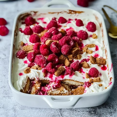 Un dessert présenté dans un plat blanc, surmonté de crème, framboises et éclats de biscuits.